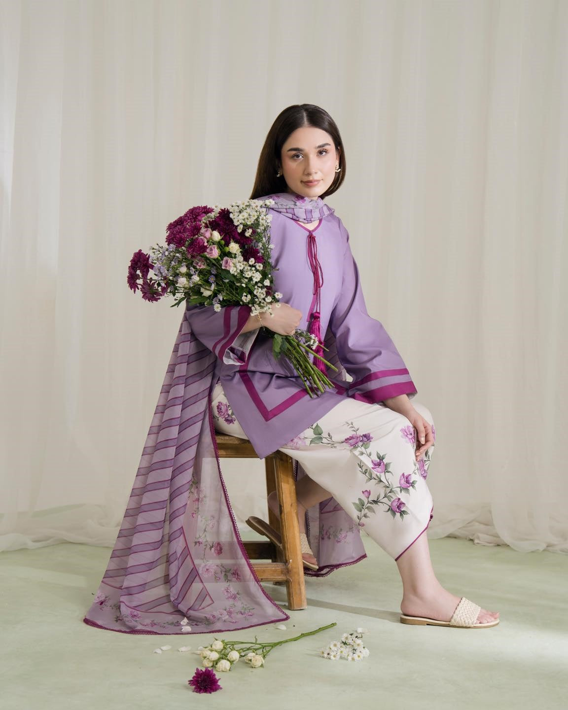 Woman in a purple outfit with floral patterns holding flowers against a white curtain background