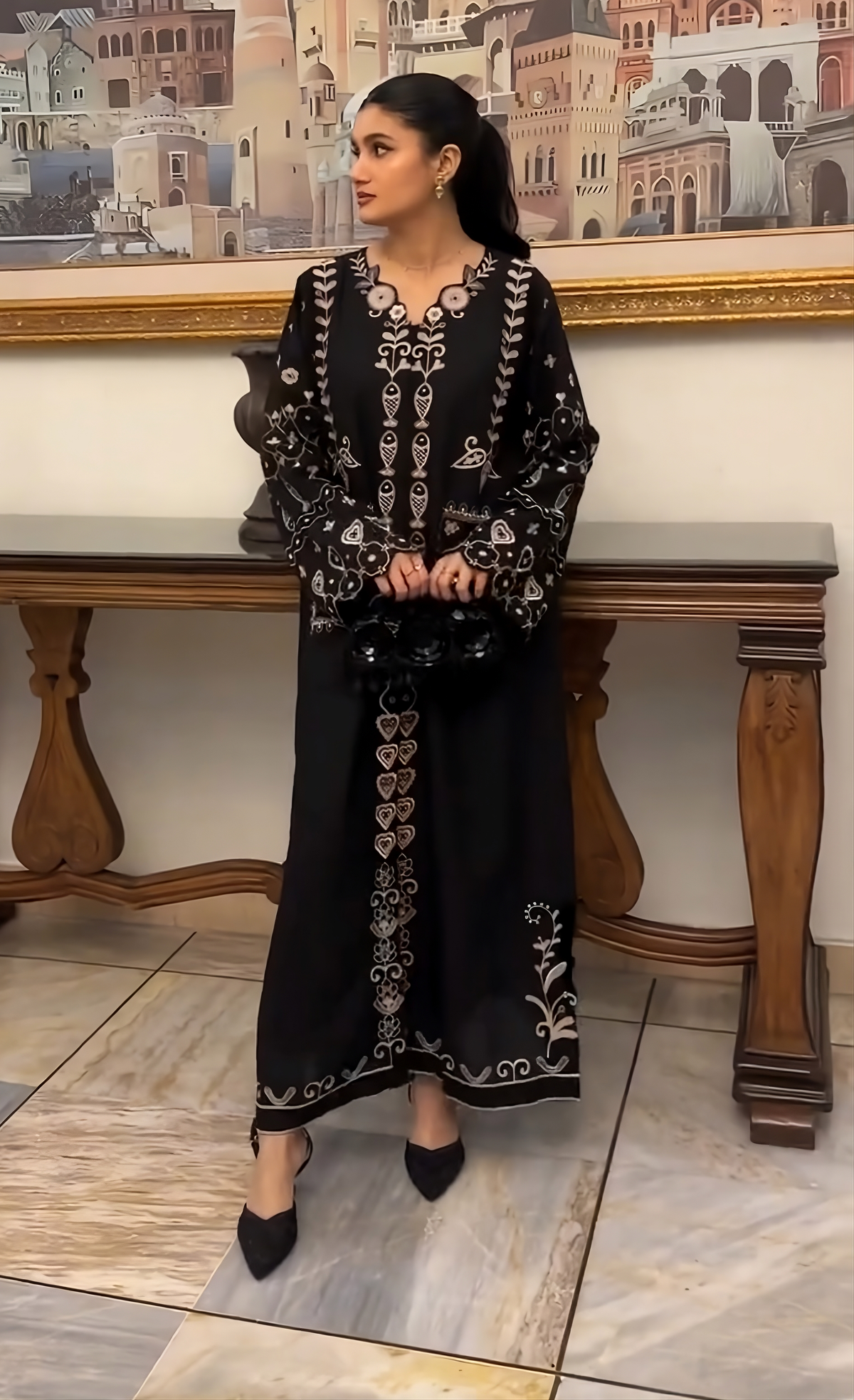 A woman models a black long-sleeve abaya dress with intricate silver and white embroidery, standing in front of a wooden console table and a framed picture.