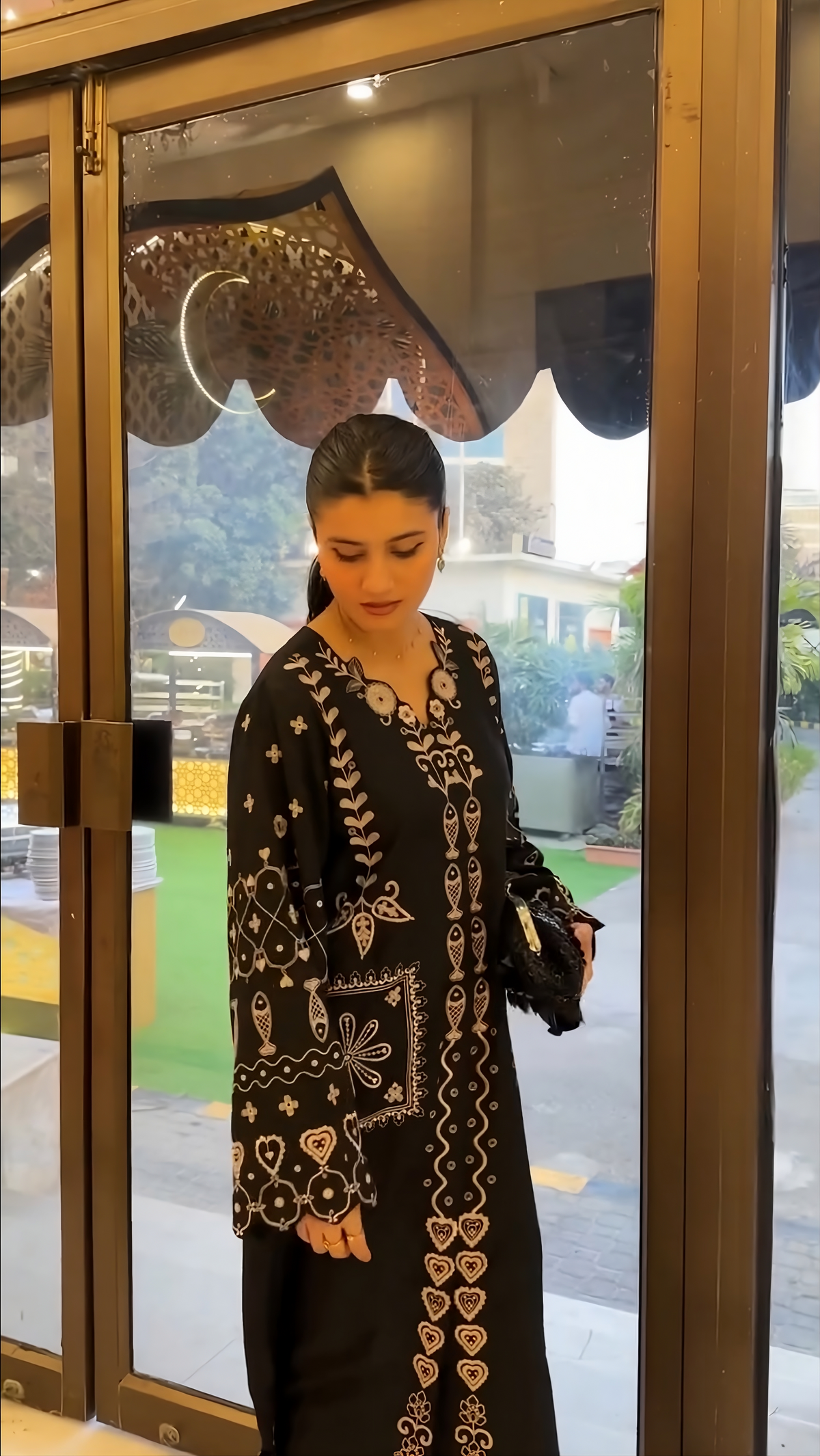 A woman wearing a black long-sleeve embroidered dress with intricate gold and white patterns, standing in front of a glass door.
