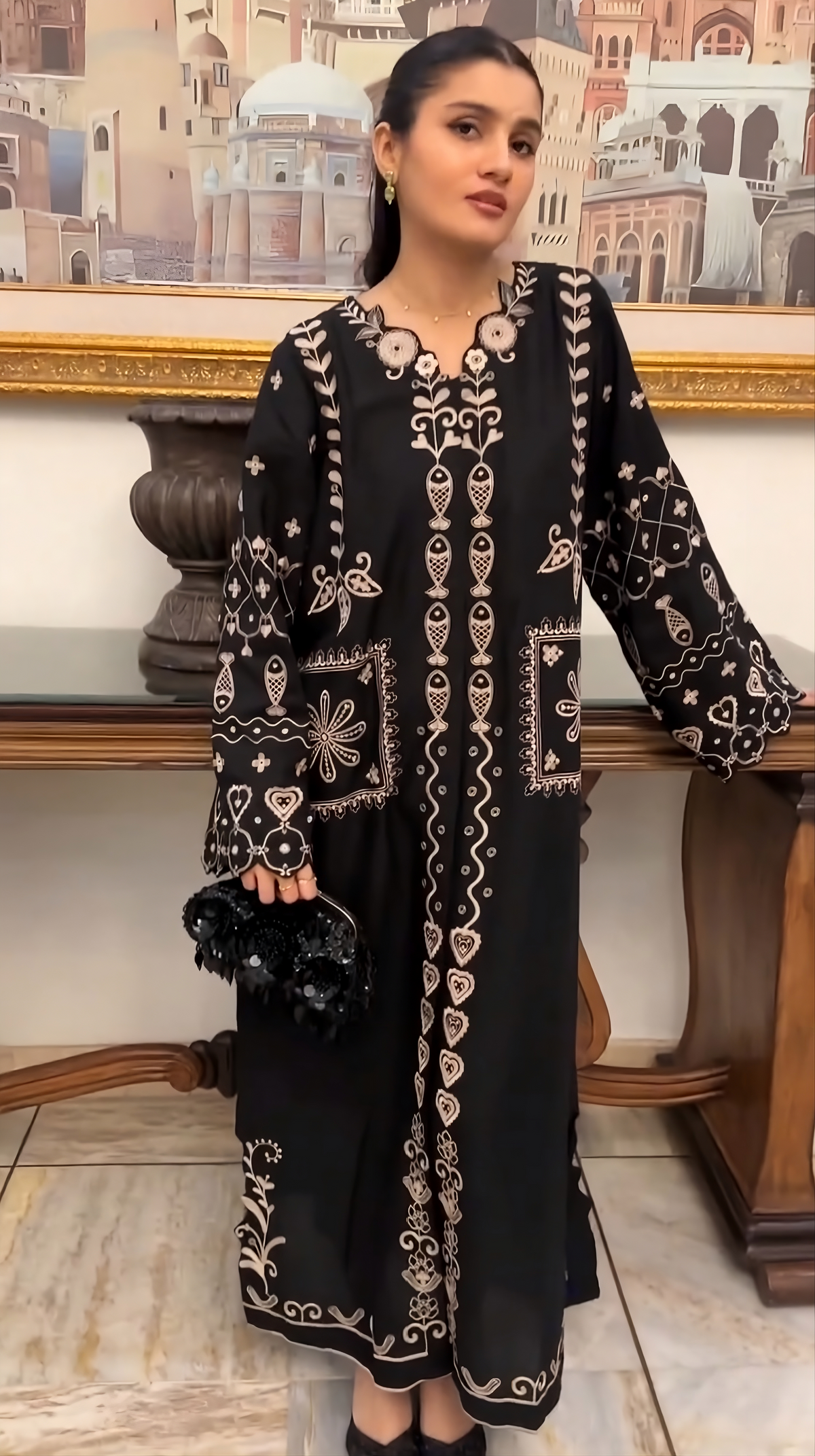 A woman modeling a long black tunic dress with intricate beige embroidery, holding a black embellished clutch in an indoor setting with a wooden console table and framed artwork in the background.