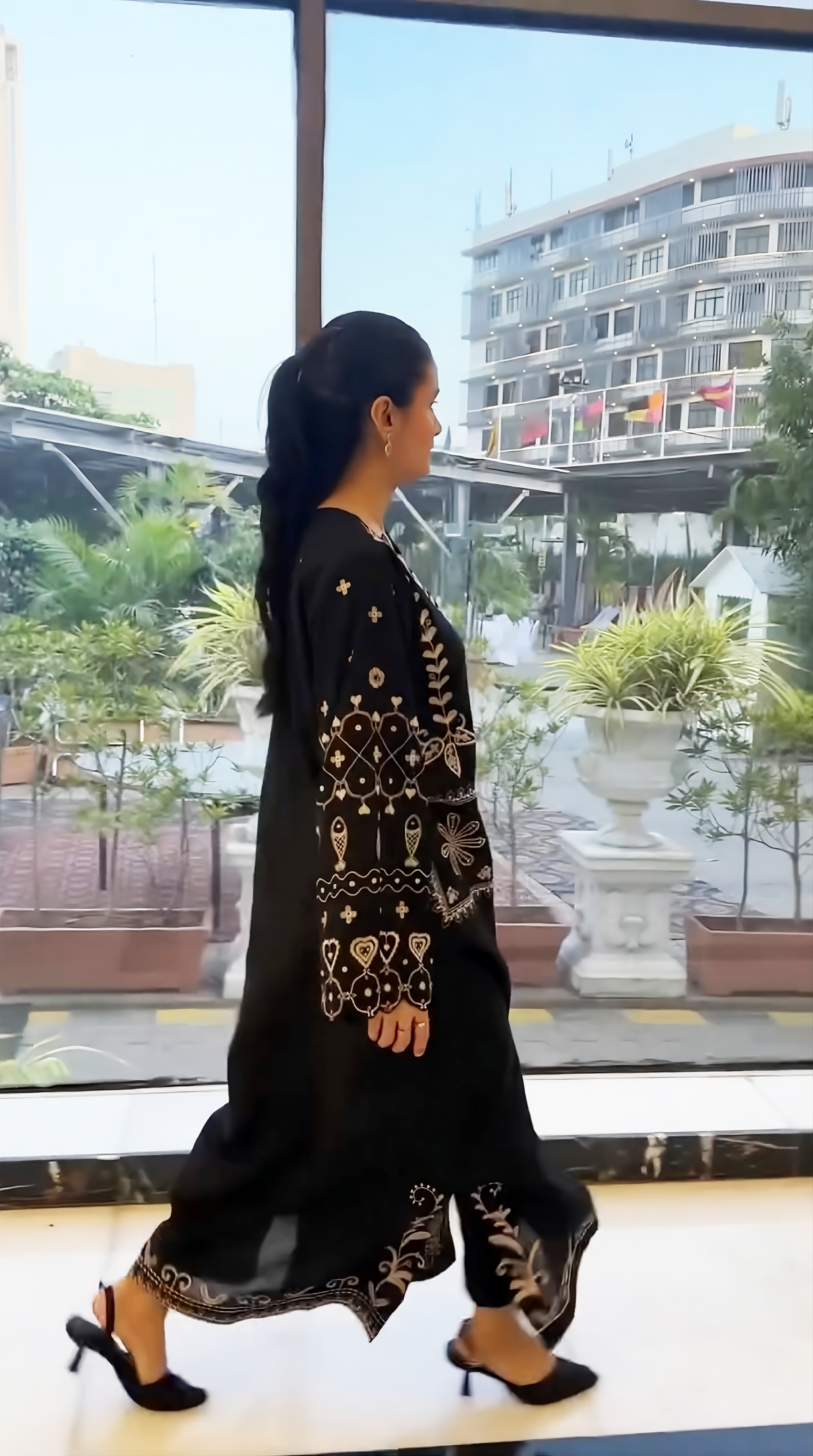 A woman wearing a black long kurta with intricate gold embroidery walks past a large window in an indoor setting with an outdoor view.